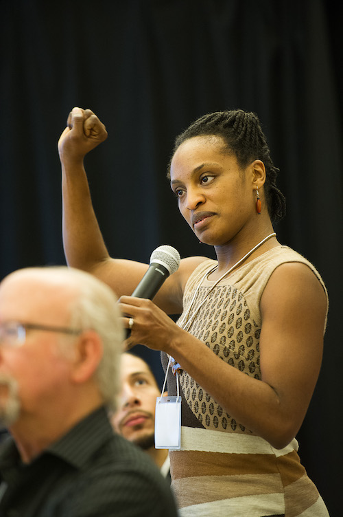 A woman in the audience stands and raises her hand in a loose fist as she speaks during the Diversity in Dance Education panel